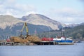 Crane lifting wood logs from stack to ship in sea loch for transportation of biomass fuel in Scotland