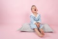 Cranky child boy refuses to go to bed. Kid boy in bathrobe sits on pillow on pink background Royalty Free Stock Photo