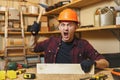 Handsome smiling young man working in carpentry workshop at wooden table place with piece of wood