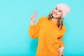 Crazy beautiful trendy girl in mustard coloured sweater and pink beanie hat making peace sign hand gesture. Cool young woman.