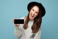 crazy beautiful young woman wearing black hat and grey sweater having fun holding phone showing smartphone isolated on Royalty Free Stock Photo