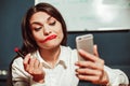 Crazy office manager businesswoman doing makeup with red lipstick at workplace using mobile phone instead of mirror Royalty Free Stock Photo