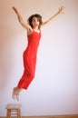 Crazy positive young brunette woman in red overalls jumping up from wooden stool at home