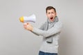 Crazy young man in gray sweater, scarf holding megaphone keeping mouth wide open on grey background. Healthy