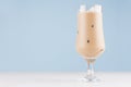 Creamy alcohol coffee cocktail with ice cubes in elegant wineglass on white table and soft light blue wall.