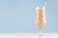 Creamy cold fresh beverage in elegant transparent glass with ice cubes and striped straw in modern light blue interior.