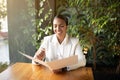 Create idea for startup. Cheerful young black woman in white suit sitting at table reading menu in cafe