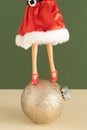 Creative Christmas layout with a doll wearing a red Santa Claus suit and hat stands on a decorative bauble against a green Royalty Free Stock Photo