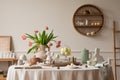 Creative composition of easter dining room interior with round table, vase with tulips, colorful eggs, pitcher, wooden trace,