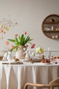 Creative composition of spring easter dining room interior with round table, vase with tulips, colorful eggs, wooden trace, bread