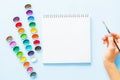 Creative flat lay of watercolor palettes, notebook, female hand holding paint brush. Artist workplace on a blue pastel background Royalty Free Stock Photo