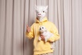 Creative lama person in yellow hoodie holding little white dog showing thumb up on beige studio wall background