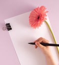 Creative Mother`s Day greeting card design. The hand of a girl writing a greeting card, blank paper and a pink gerbera flower on a Royalty Free Stock Photo