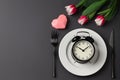 Creative table setting with alarm clock on a white plate, black fork and knife, tulips on a dark background. Breakfast, Wedding