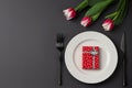 Creative table setting with gift box on a white plate, black fork and knife, tulips on a dark background. Valentine`s Day, Weddin