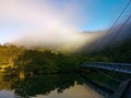 Beautiful view of creek with reflections blue sky, light clouds, fog, suspension bridge, mountains and trees on water Royalty Free Stock Photo