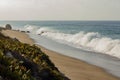 Cresting breaking wave on shoreline edge with washback foam on sandy shoreline with boulders