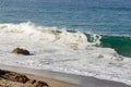 Cresting breaking wave on shoreline edge with washback foam on sandy shoreline with boulders