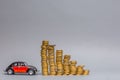 Crimson car figurine sitting next to a row of decreasing in size columns of coins, on gray background Royalty Free Stock Photo