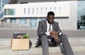 Crisis and unemployment concept. Upset black office worker sitting with box of personal stuff on stairs of office center Royalty Free Stock Photo