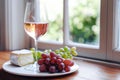 A crisp glass of rose wine sits on a windowsill, accompanied by a plate with a selection of cheese and ripe grapes. Royalty Free Stock Photo