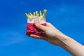 Crispy french fries in a paper bag in a male hand on a blue sky background.