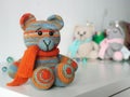 Crochet bear is orange-blue in scarf on background of other toys on light background. Knitted soft toy Teddy