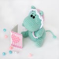 Crocheted amigurumi toy small turquoise hippo in a pink-white cap with a sad surprised look reads a book with a heart on cover