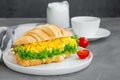 Croissant with scrambled eggs, lettuce and cherry tomatoes on a white plate with a cup of coffee on a dark concrete background. Royalty Free Stock Photo