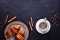 Croissants on a ceramic plate, a cup of coffee, cinnamon sticks and coffee beans on a black wooden table.