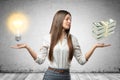 Crop image of young beautiful businesswoman, hands at sides, palms facing up and levitating lightbulb and stack of cash. Royalty Free Stock Photo