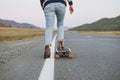 Crop photo of women`s legs in jeans on longboard on road with the mountains background