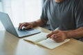 Crop picture of hand freelance man using pen writing notes notebook and working on laptop on table at home. Entrepreneur man