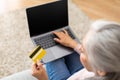 Cropped european senior woman with gray hair with credit card and laptop shopping online in room interior