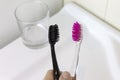 Cropped hand holding two toothbrushes of pink and black color on background of white washbowl and clear glass in bathroom.