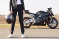 Cropped horizontal shot of female biker dressed in black leather jacket, trousers and white sneakers, holds helmet, stands on asph