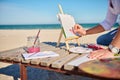 Cropped image of artist`s hand holding paintbrush and painting boat with watercolors, sitting on a wooden chaise lounge with sea Royalty Free Stock Photo