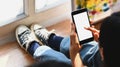 Cropped image of artist woman legs with jeans sitting on wooden floor while holding white blank screen smartphone in hands. Royalty Free Stock Photo