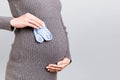 Cropped image of blue socks for a baby boy in pregnant woman`s hand against her belly at gray background. Waiting for a future