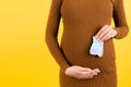 Cropped image of blue socks for a baby boy in pregnant woman`s hand against her belly at yellow background. Waiting for a future