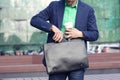 Cropped image businessman in casual wear with cup of coffee to go puts smartphone into trendy leather briefcase against modern