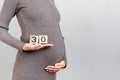 Cropped image of cubes with thirty weeks of pregnancy in pregnant woman`s hands wearing gray dress at gray background. Upcoming
