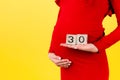 Cropped image of cubes with thirty weeks of pregnancy in pregnant woman`s hands wearing red dress at yellow background. Upcoming