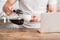 cropped image of man pouring coffee from coffee pot into cup near laptop