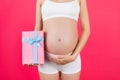 Cropped image of pink spotted gift box in pregnant woman`s hand against her belly at pink background. Future mother in white