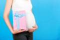 Cropped image of pink spotted gift box in pregnant woman`s hand against her belly at blue background. Future mother in home