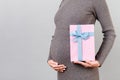 Cropped image of pink spotted gift box in pregnant woman`s hand against her belly at gray background. Future mother in gray dress