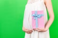 Cropped image of pink spotted gift box in pregnant woman`s hand against her belly at green background. Future mother in white