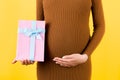 Cropped image of pink spotted gift box in pregnant woman`s hand against her belly at yellow background. Future mother in brown