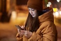 Cropped image of pleasant looking woman concentrated in smart phone, dressed in stylish headgear and jacket, has outdoor walk in Royalty Free Stock Photo